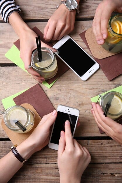 A close-up shot of a diverse group of hands pointing at a smartphone displaying a coupon code on a wooden table during a meeting. The background suggests a relaxed coffee shop setting.