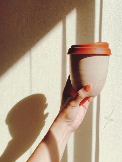A minimalist image showing a hand holding a reusable coffee cup next to a plant, with a blurred background of a street market emphasizing local and sustainable commerce. The focus is on eco-friendly choices.