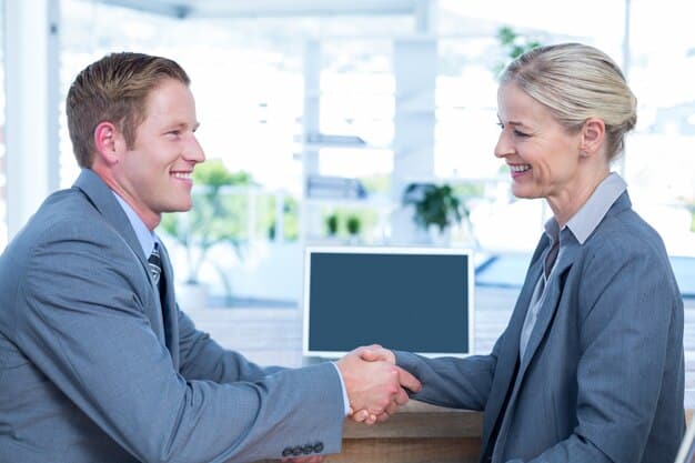 Two business people shaking hands across a polished conference table, with laptops and documents, signifying a successful agreement and collaboration.