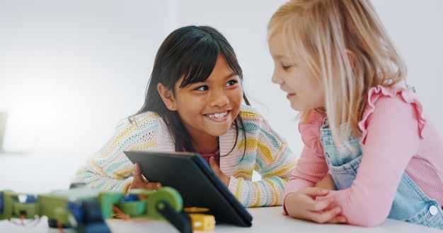 A teacher helping two smiling elementary school students work on a coding project on a tablet in a modern classroom.