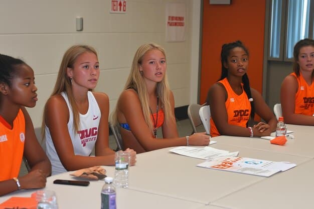 A group of diverse college athletes in various sports uniforms gathered around a table with a professional agent, reviewing endorsement contracts.