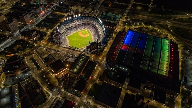 Aerial view of a bustling baseball stadium during a night game. The field is brightly lit, with players in action. The stands are packed with fans, creating a sea of colors from team jerseys. Fireworks explode in the background, adding to the festive atmosphere. This image represents the high attendance success and excitement seen in some teams contributing to MLB Attendance Up 8% This Season: Is Baseball Making a Comeback?
